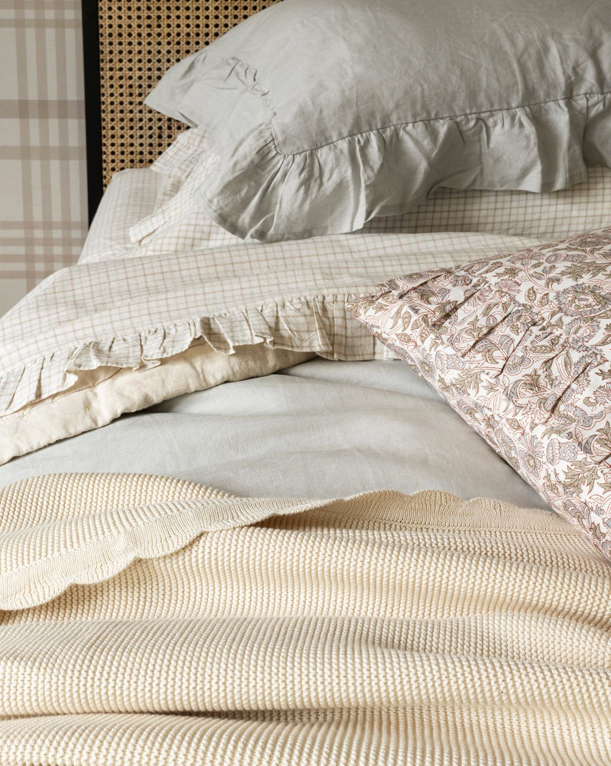 A neatly made bed features the Pinnacle Knit Kids Blanket, a textured 100% cotton layer, paired with ruffled light gray and beige pillows, a paisley-patterned pillow, and a plaid sheet for a cozy, inviting look.