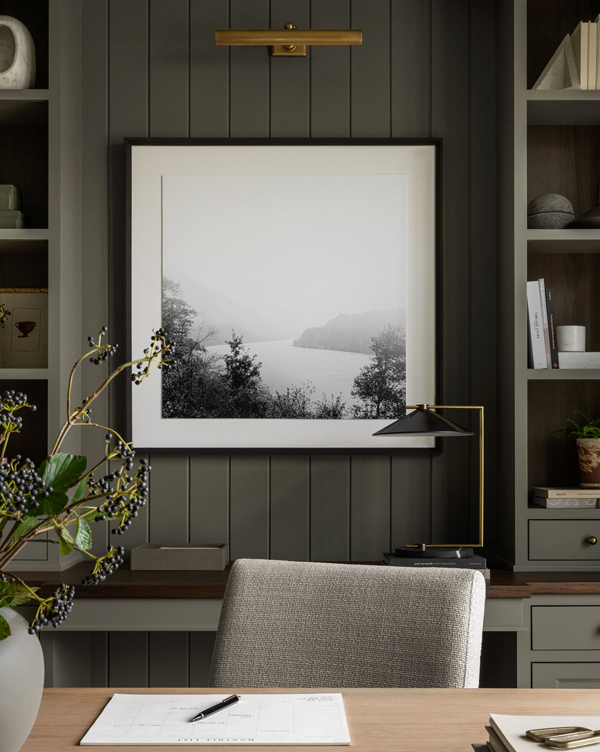 A modern home office with a gray panel wall, a framed Mist on the Lake black and white photography print, a desk with papers and pen, a gray upholstered chair, and built-in shelves filled with books and decor.