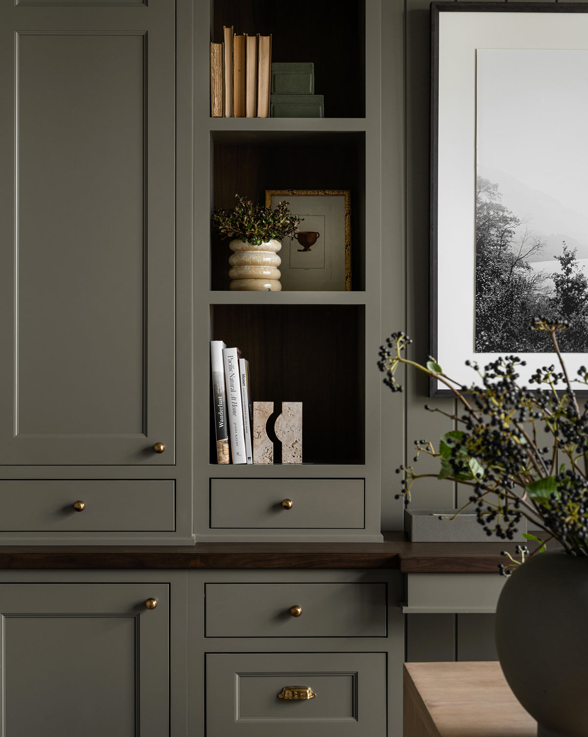 Built-in shelves and sage green cabinets are styled with books, a leafy plant in a ceramic vase, the Travertine Object as decor, framed art, and brass handles. A black-and-white photo hangs on the wall.
