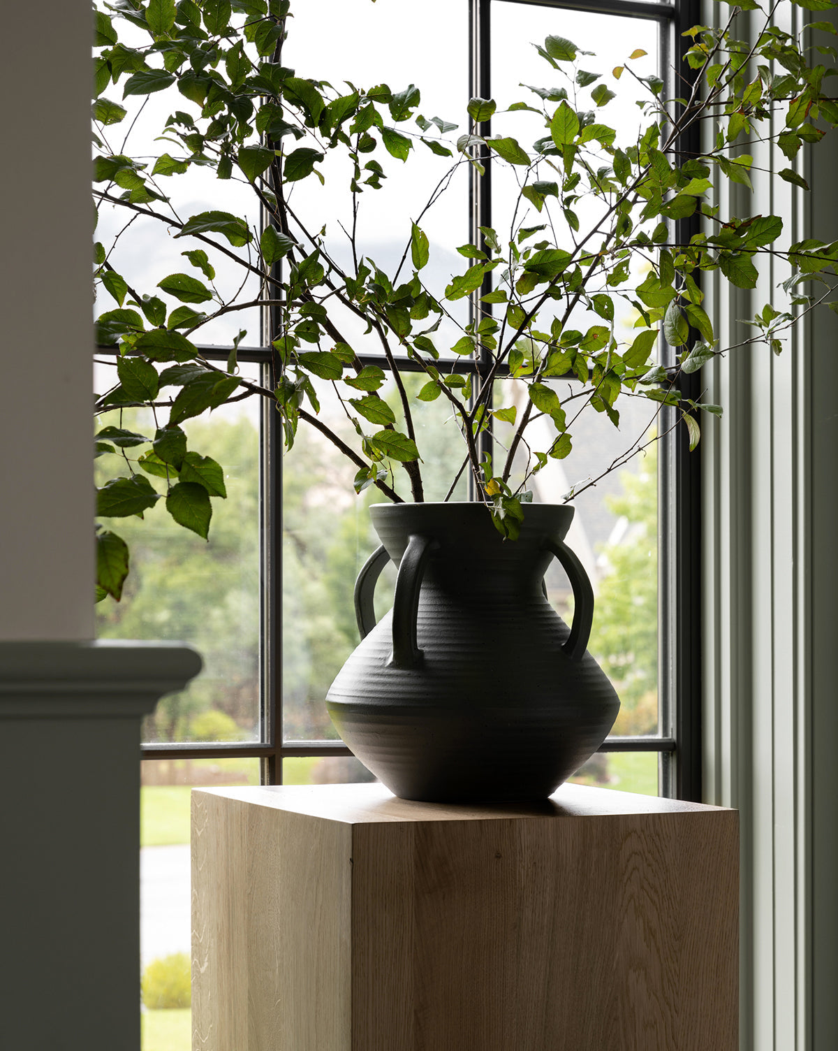 The Isaiah Vase, featuring a unique black ceramic silhouette and leafy branches, is displayed on a light wooden pedestal in front of a large window overlooking lush greenery and trees.