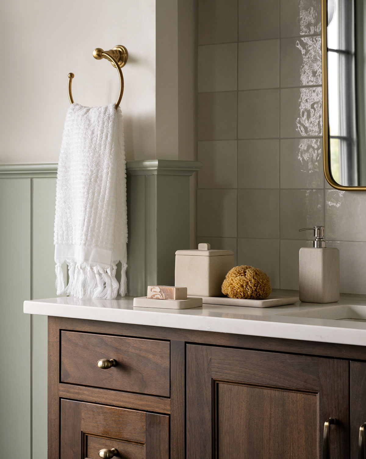 A dark wood vanity and green wall set an organic tone, while the Amada Sandstone Bath Collection—with soap dispenser, bar soap, sponge, and small container—adorns the countertop. A gold ring holds a white towel above.