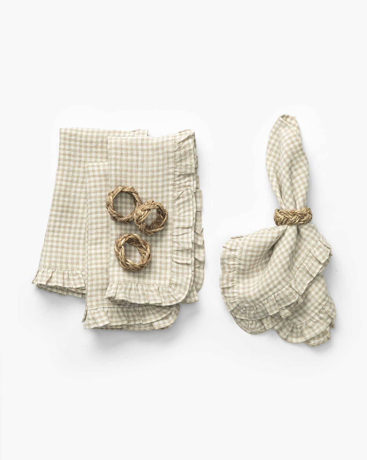 The Catania Linen Napkins (Set of 4) in beige and white gingham are styled with ruffled edges—three folded with braided straw napkin rings, and one elegantly unfolded—displayed against a white background, by McGee & Co.