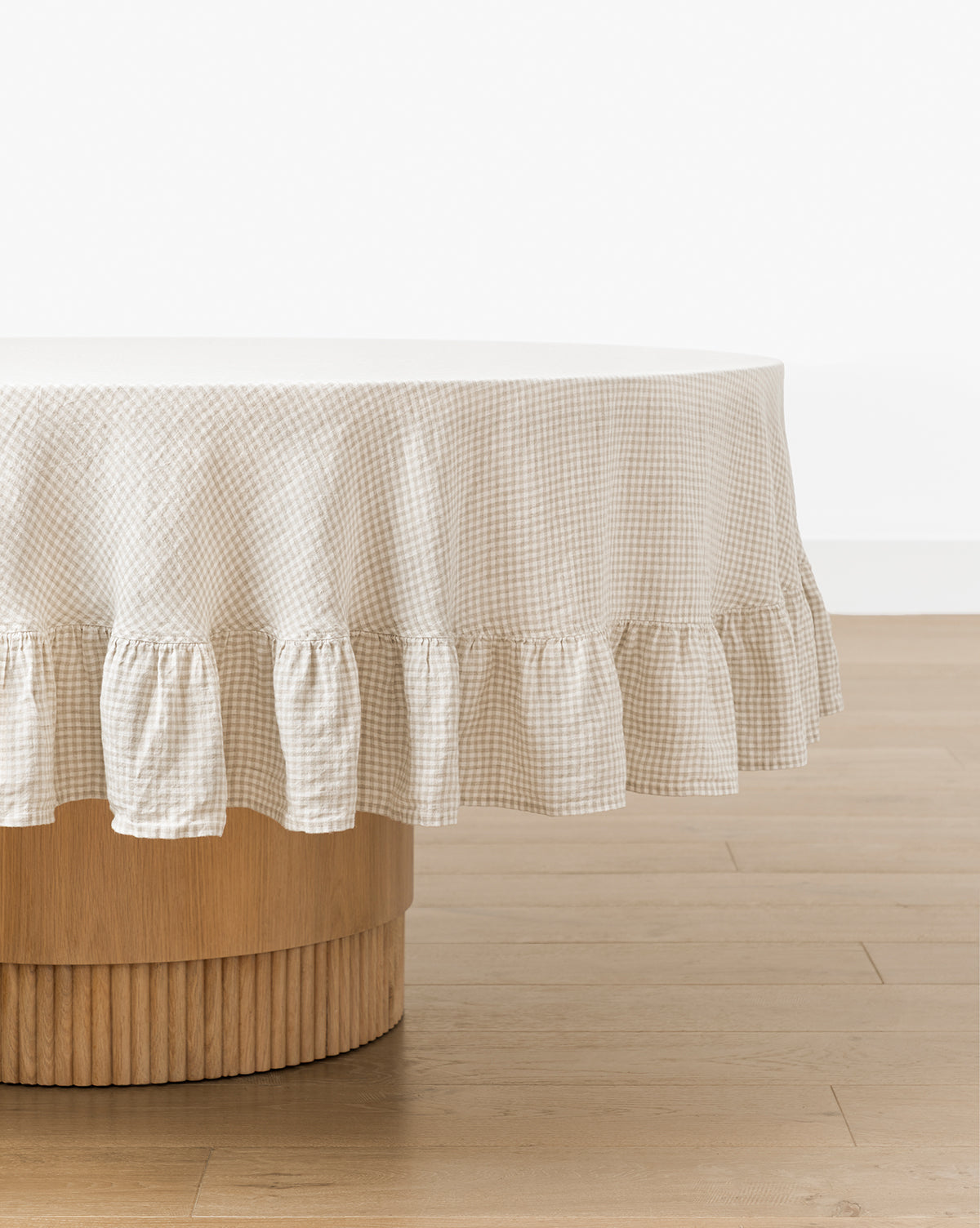 A round wooden table is covered with the Catania Linen Ruffle Tablecloth in beige and white gingham, set on a light wood floor against a plain white wall, by McGee & Co.
