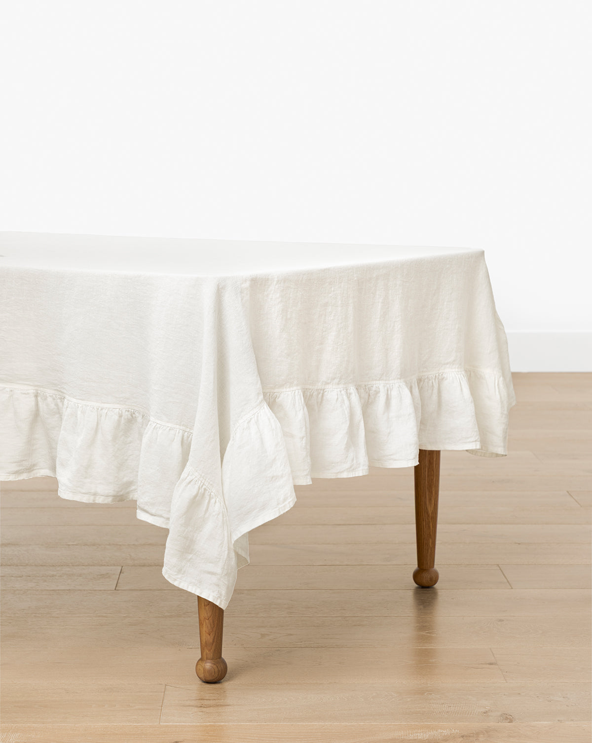 A rectangular wooden table with curved legs is draped in the Catania Linen Ruffle Tablecloth, by McGee & Co. The table stands on a light wood floor against a plain white wall.