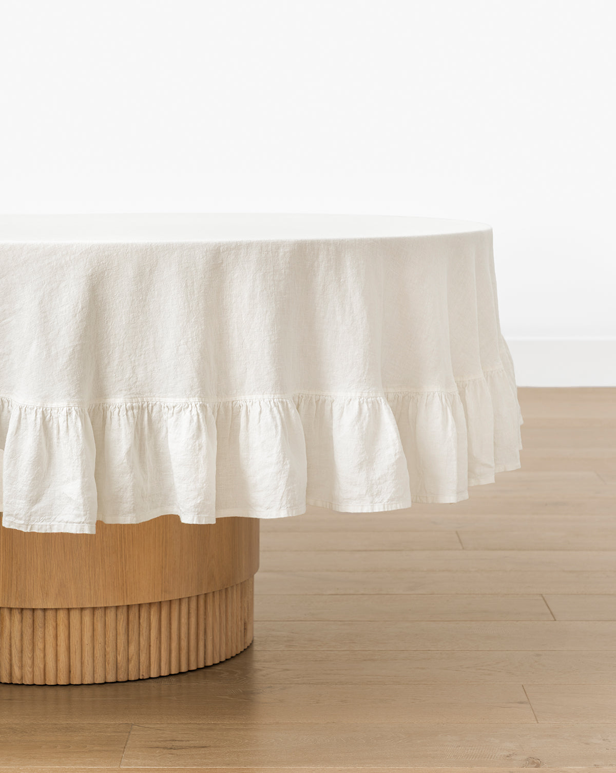 The Catania Linen Ruffle Tablecloth, with a delicate ruffled edge, covers a round wooden table with a ribbed base, displayed on a light wooden floor against a plain white wall, by McGee & Co.