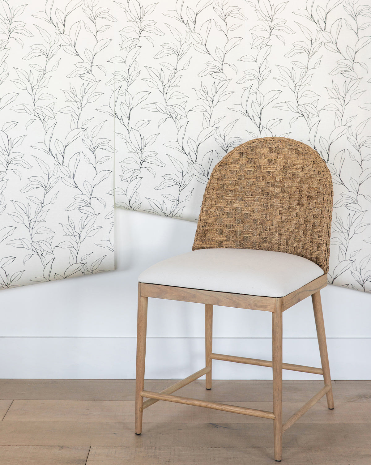 A wooden chair with a woven rattan backrest and cream cushion sits before a white wall featuring Cecile Floral Print Wallpaper by York Wallcoverings, its light leafy pattern partially peeling at the bottom—a vignette that feels straight from McGee & Co.