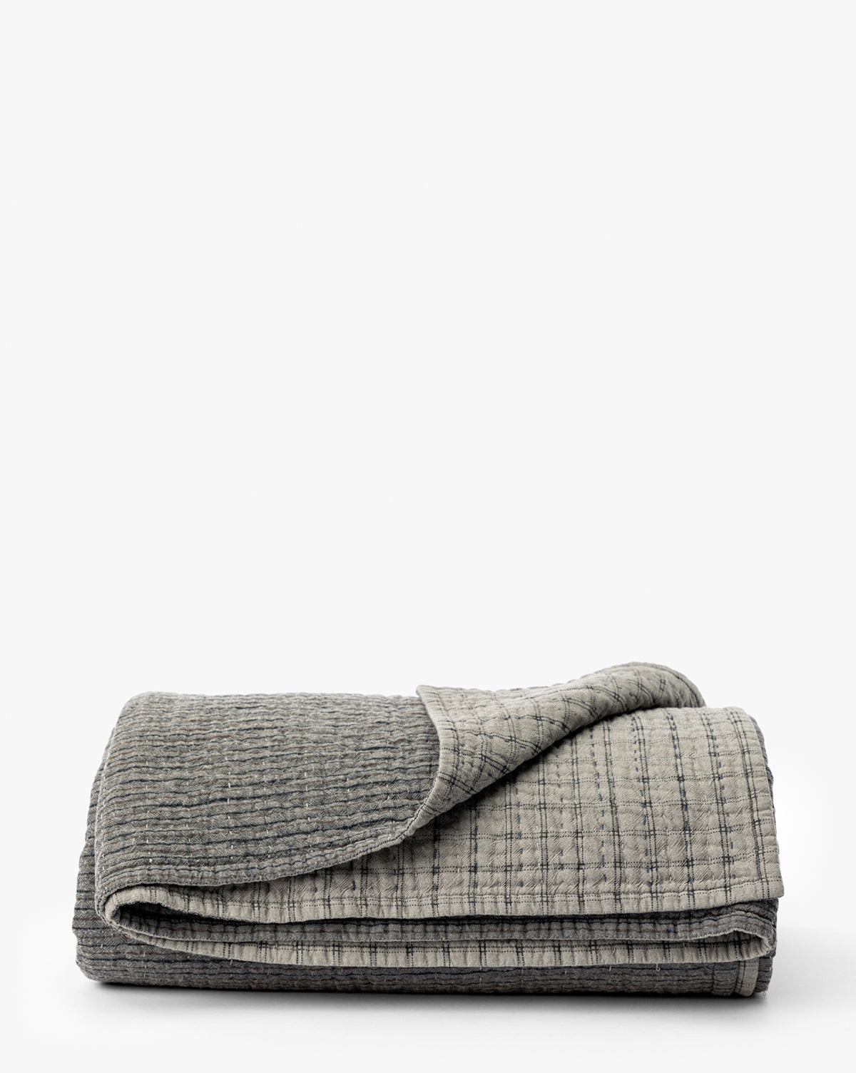 The Cecily Kids Quilt by Renuka Inc. is a textured gray blanket with a visible stitched pattern, neatly folded on a flat white surface. One corner is slightly flipped over to show the underside, McGee & Co.