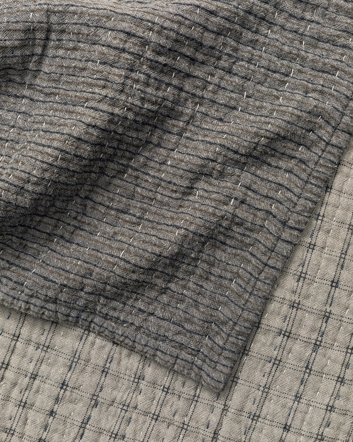 A close-up of two overlapping Cecily Kids Quilts by Renuka Inc. shows textured, slightly wrinkled fabrics: the top features a darker grid pattern with fine lines, while the bottom displays a lighter shade with bold grid lines—McGee & Co.