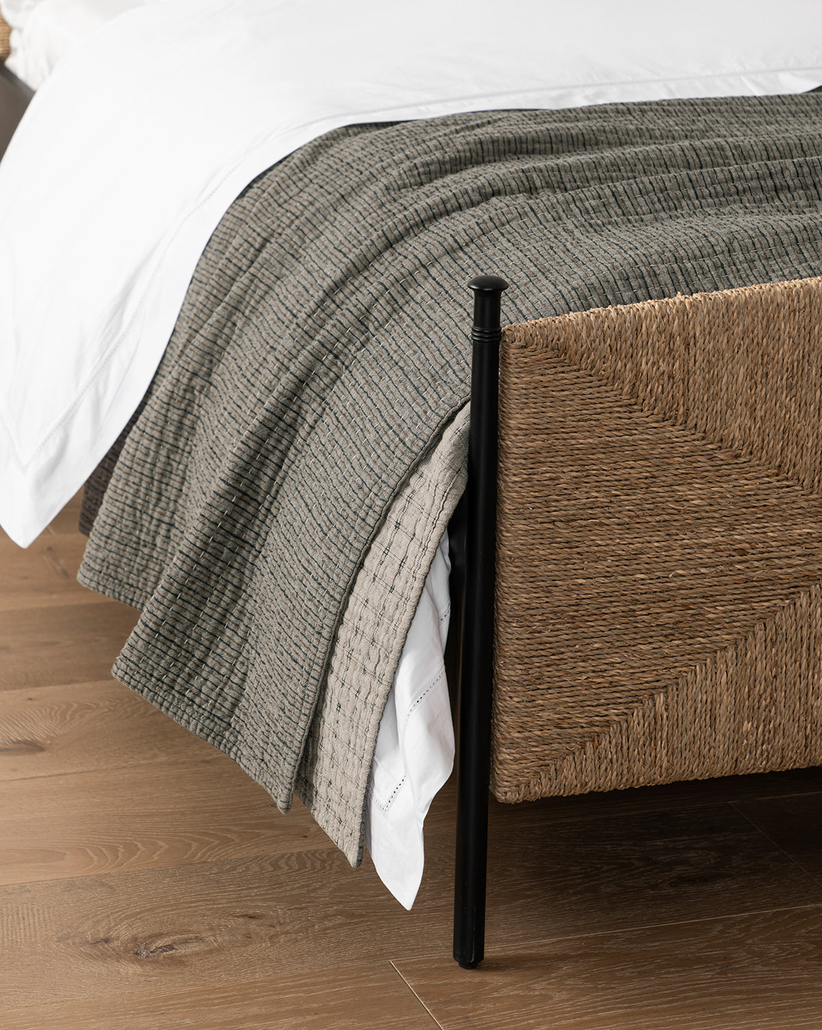 A close-up of a bed corner with a woven rattan frame, white sheet, Cecily Kids Quilt, gray textured blanket, and a patterned beige throw, all on a wooden floor by McGee & Co.