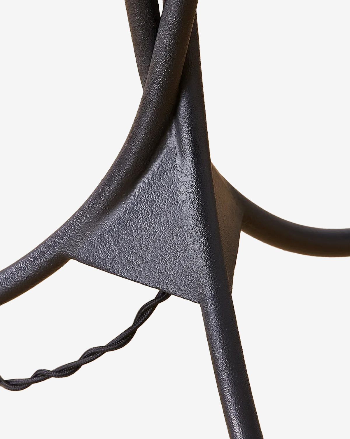 Close-up of the Cedar Floor Lamp’s intersecting forged iron rods with a textured finish, joined by a triangular metal plate, and featuring a twisted black electrical cord running through the structure, by McGee & Co.