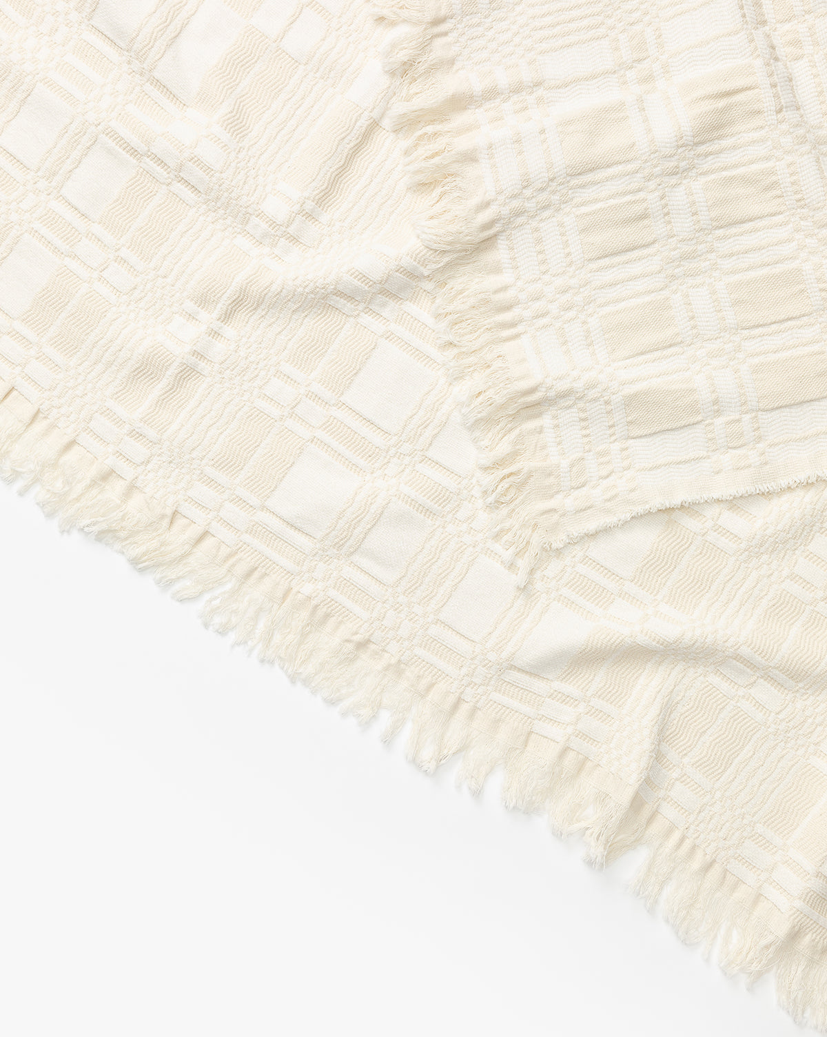 A close-up of the Chamberlain Coverlet by Makerspalm, featuring cream-colored cotton with a textured plaid pattern and fringed edges, arranged diagonally on a white background by McGee & Co.