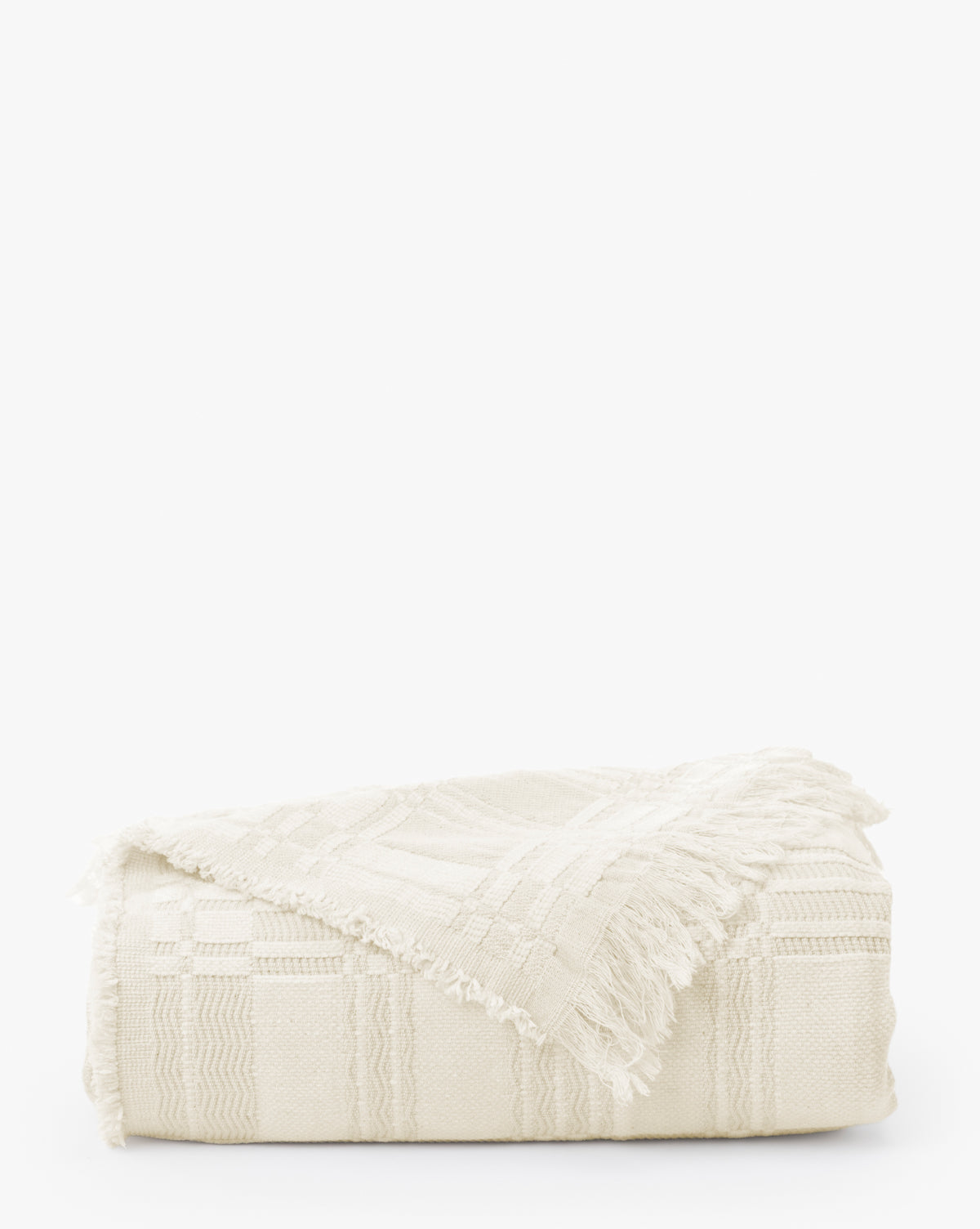 The Makerspalm Chamberlain Coverlet is a folded off-white woven blanket with a subtle geometric pattern and fringed edges, displayed on a plain white background. McGee & Co.