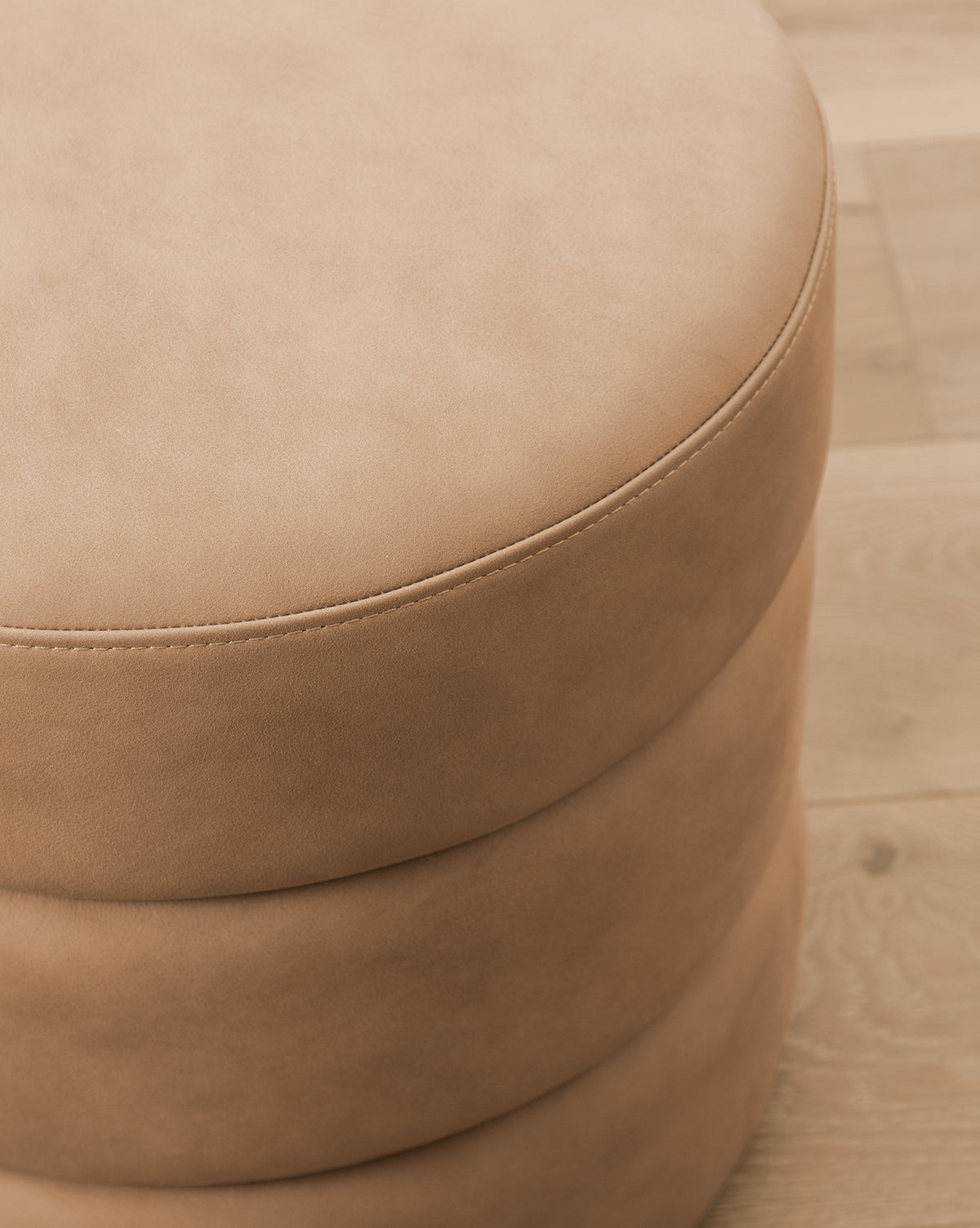 Close-up of the Channel Ottoman, a round, tan piece featuring visible top stitching and subtle quilted details, placed on a light wood floor to bring midcentury charm to any room, by McGee & Co.