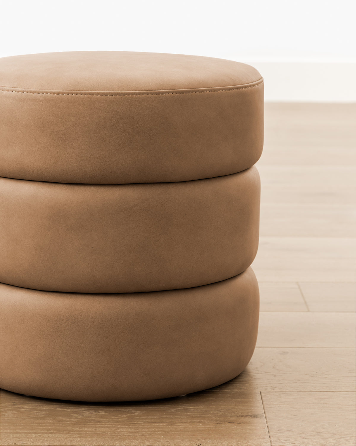 A close-up of the Channel Ottoman, a round, light brown, three-tiered piece with smooth upholstery, sits on a light wood floor against a white wall, bringing midcentury flair to the room by McGee & Co.