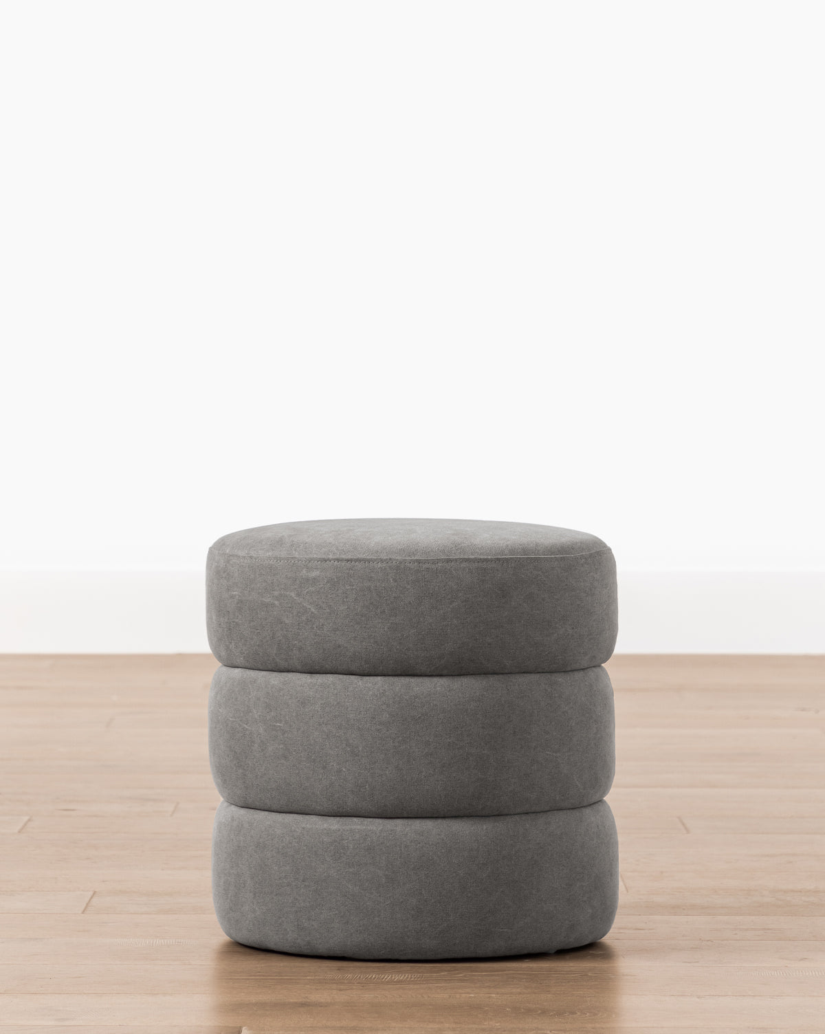 The Channel Ottoman features a round, gray design with three stacked cushion-like layers and a subtle quilted texture, adding midcentury charm as it sits on a light wood floor against a plain white wall, by McGee & Co.