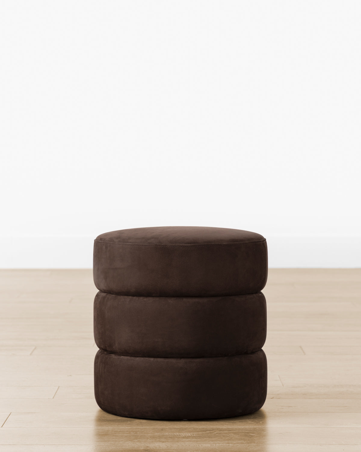 The Channel Ottoman features three stacked, quilted layers in round, dark brown upholstery, offering a subtle midcentury accent on a light wood floor against a white wall, by McGee & Co.