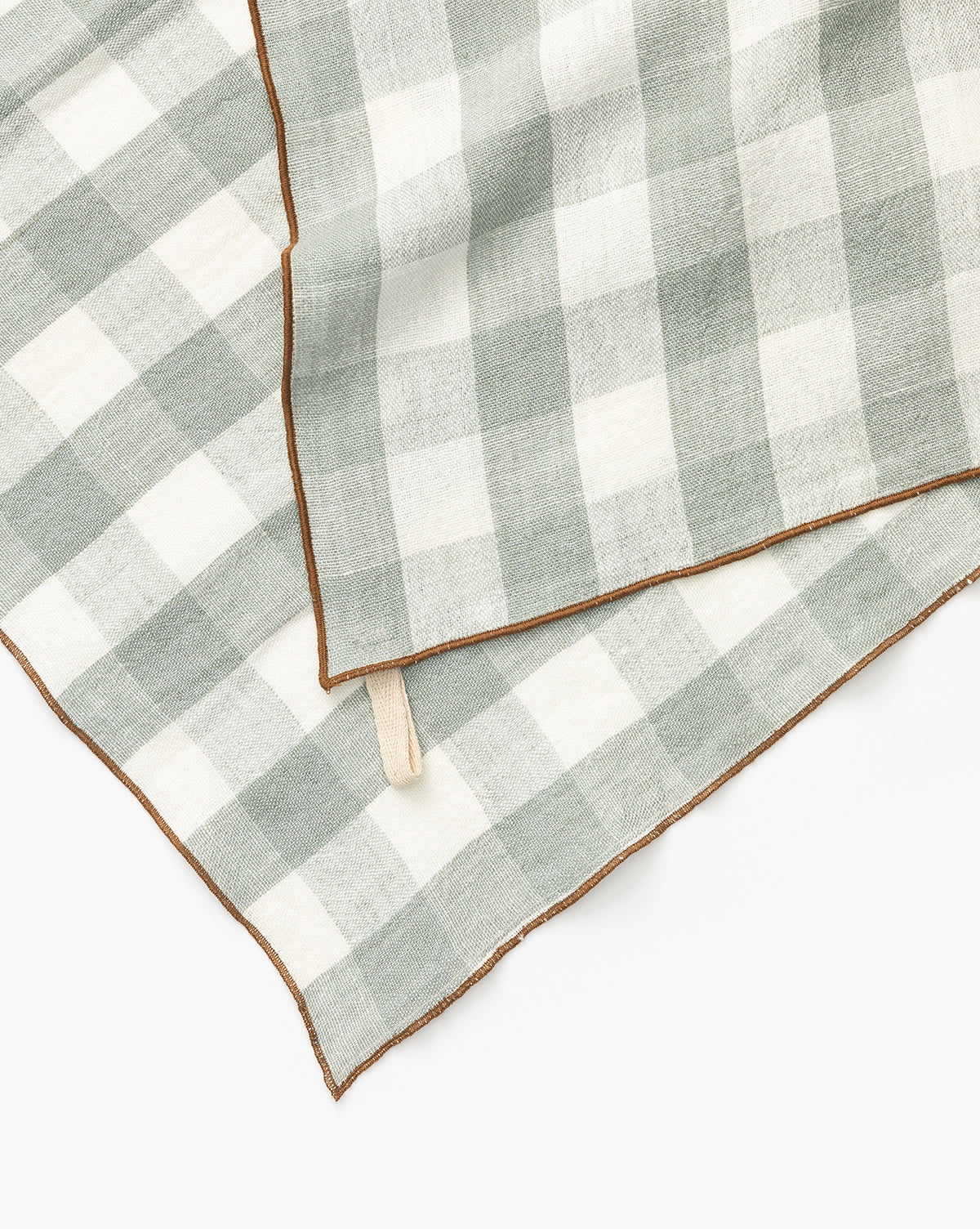 A close-up of two Charlie Check Tea Towels, featuring gray and white checkered linen with brown stitched edges, slightly overlapping on a white background—ideal for stylish table settings, by McGee & Co.