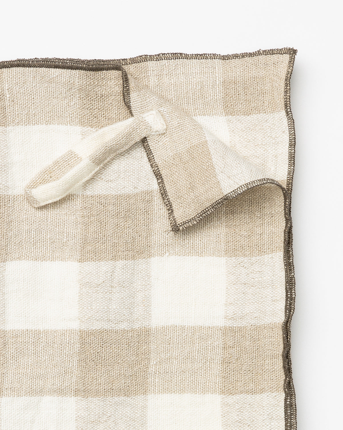 The Charlie Check Tea Towel features a beige and white checkered linen design with a hanging loop, shown partially folded at one corner—ideal for elegant table settings against a white background, by McGee & Co.