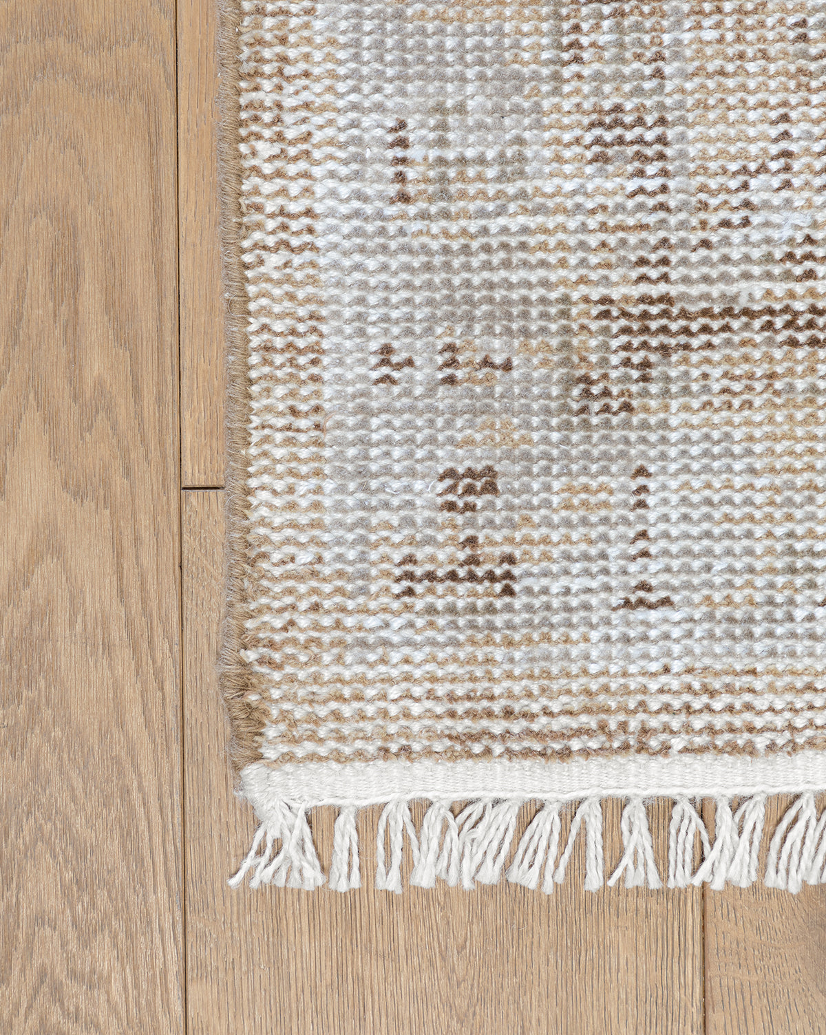 A close-up of the Charlot Hand-Knotted Rug’s corner shows its white fringe and textured beige, brown, and white pattern on a light wooden floor, by McGee & Co.