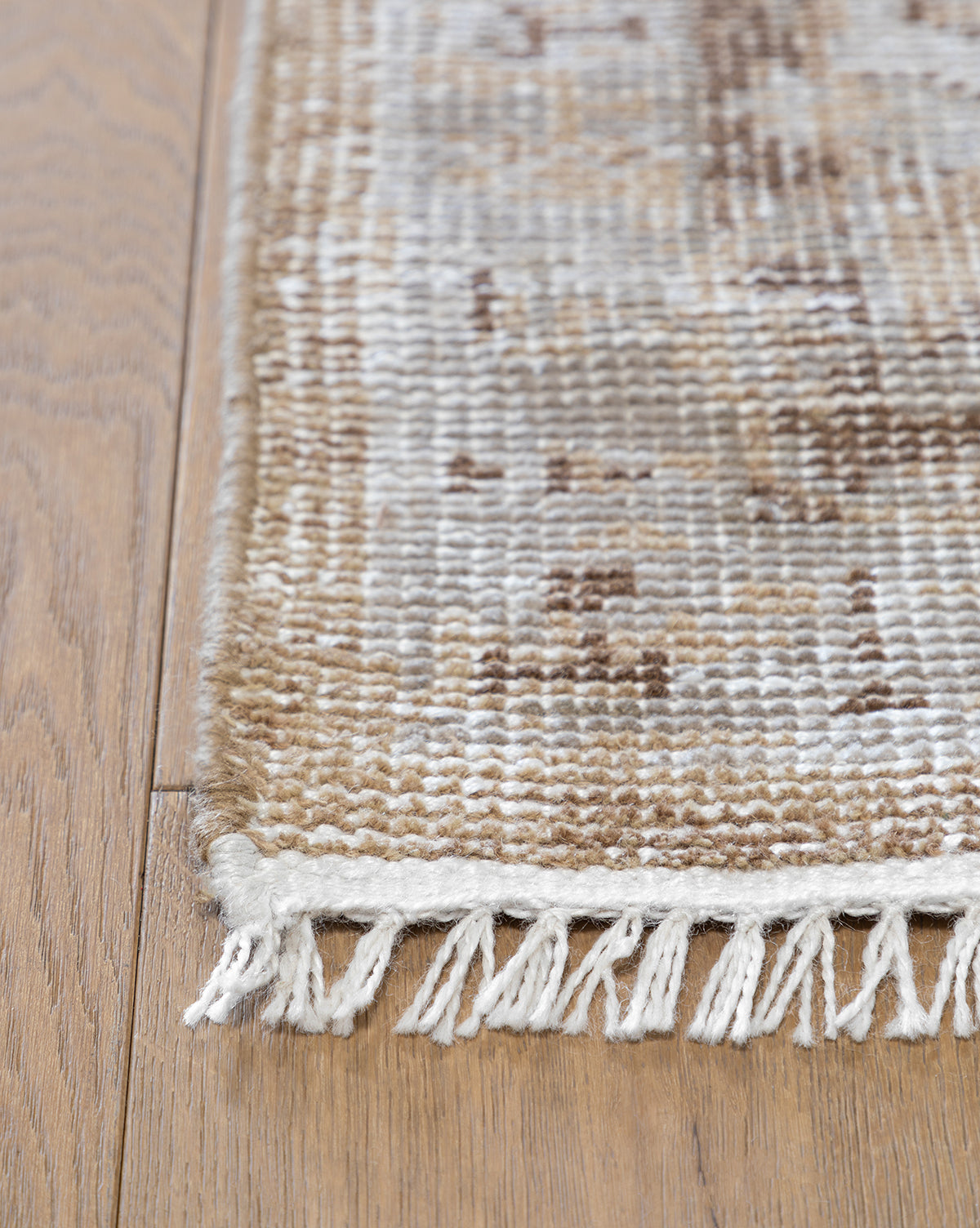 A close-up shows the corner of a Charlot Hand-Knotted Rug in beige, brown, and cream with white fringe, placed on a light wood floor, by McGee & Co.
