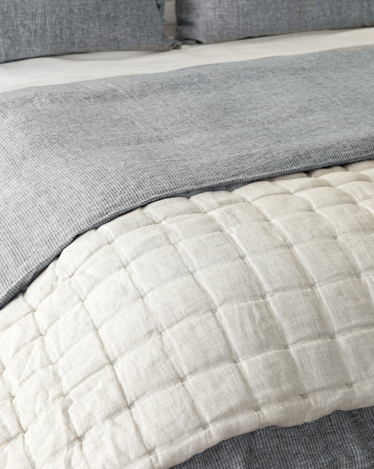 Close-up of a neatly made bed featuring the Chaucer Quilt in light beige with quilted stitching, layered with a textured gray blanket and matching gray pillows in the background.