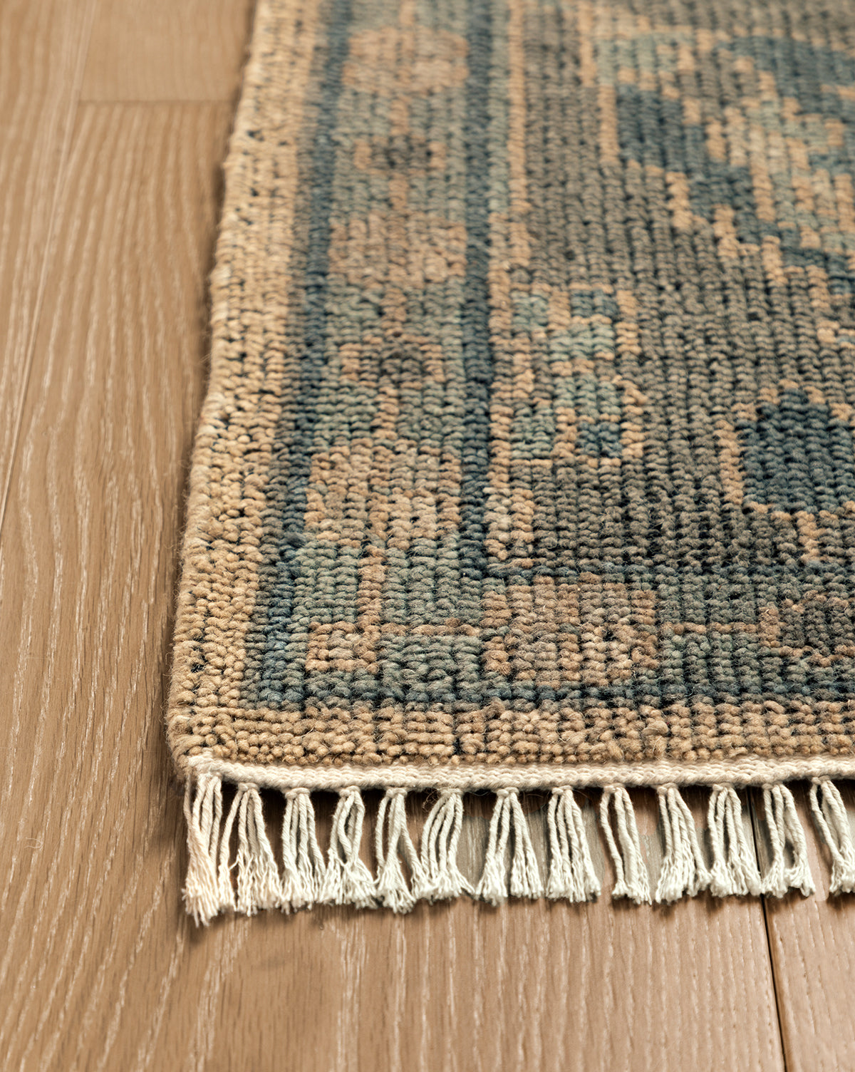 A close-up of the Chevalier Hand-Knotted Wool Rug displays its blue and beige pattern, fringed edges, and timeless elegance as it rests on light brown wooden flooring, by McGee & Co.