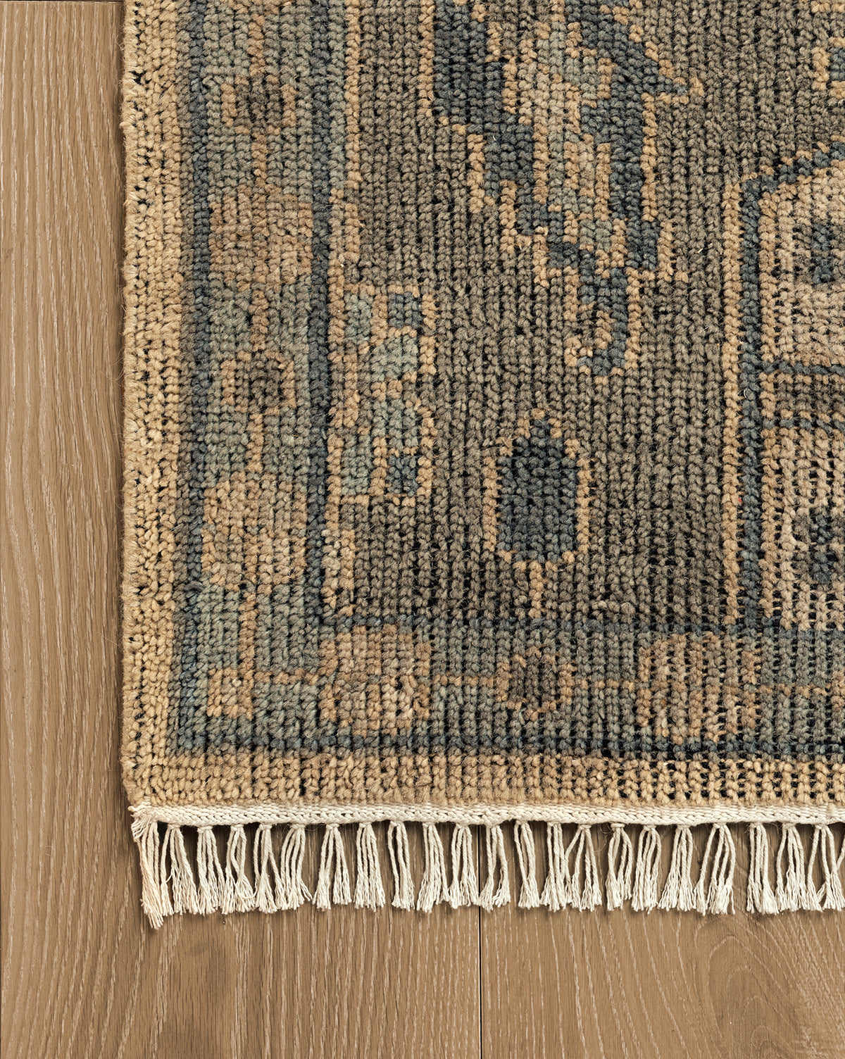 A close-up of the Chevalier Hand-Knotted Wool Rug, featuring a blue background with tan geometric patterns and beige fringe, resting on a light brown wooden floor, showcases its timeless elegance, by McGee & Co.