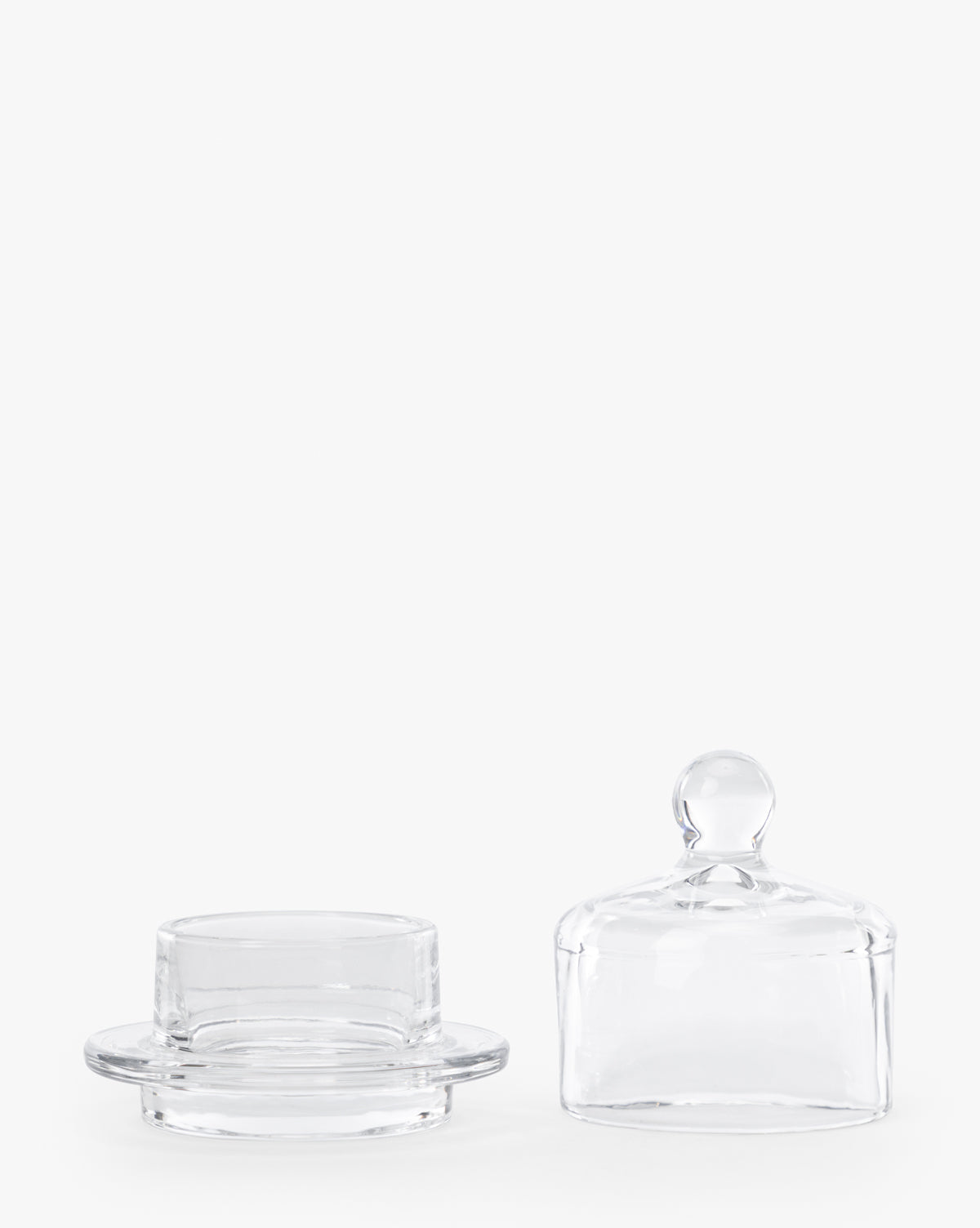 The Christian Glass Condiment Dish features a clear round base and a matching domed lid with a small knob handle, shown side by side on a white background—ideal for serving condiments or butter in style, by McGee & Co.