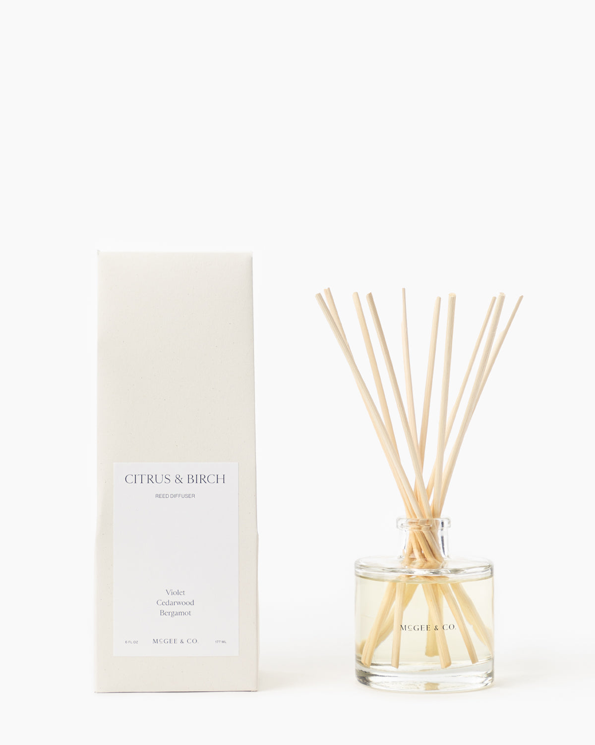 The Citrus & Birch Diffuser, a clear glass vessel with light wooden reeds, stands beside its rectangular white box labeled Citrus & Birch, elegantly enhancing your space, by McGee & Co. Both items are displayed on a plain white background.