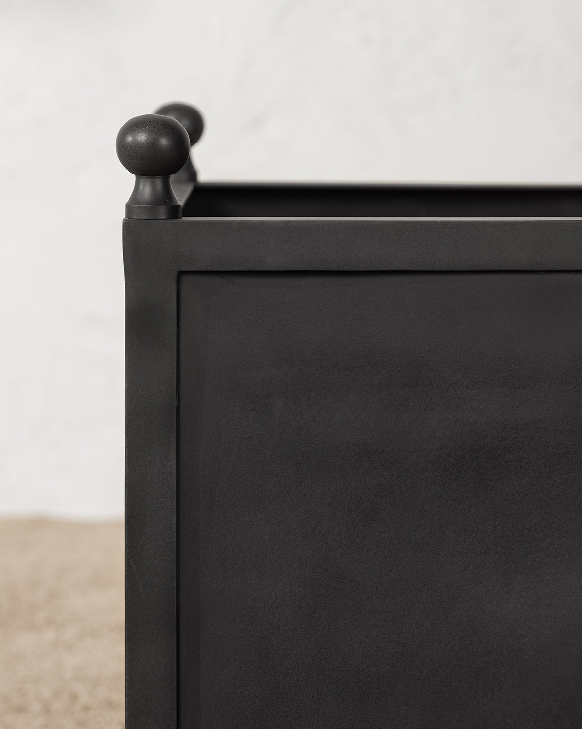 Close-up of the Clariance Planter, a black rectangular metal design with rounded finials, set against a textured white wall and pebbled surface—ideal for refined outdoor styling.
