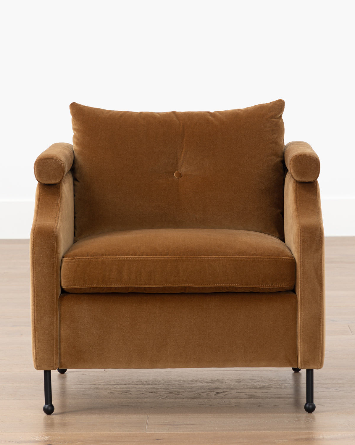 The Clegg Lounge Chair (Ready to Ship) features plush brown cotton velvet upholstery, a back cushion, rounded armrests, and sturdy iron legs, set on a light wood floor against a plain white wall by McGee & Co.
