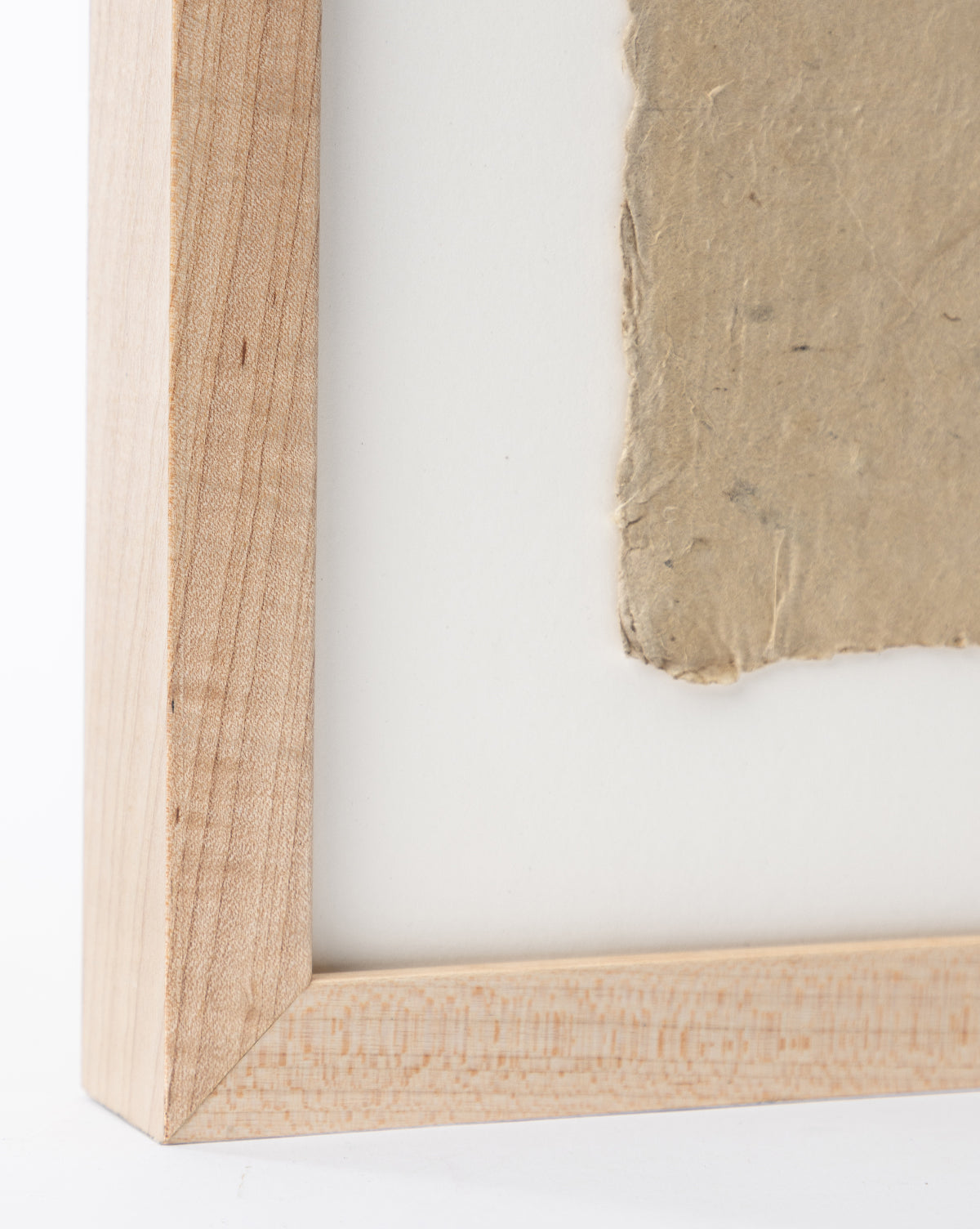 Close-up of a maple wood frame’s mitered corner, featuring Collage 1, handmade paper art with rough edges, mounted on a white background.