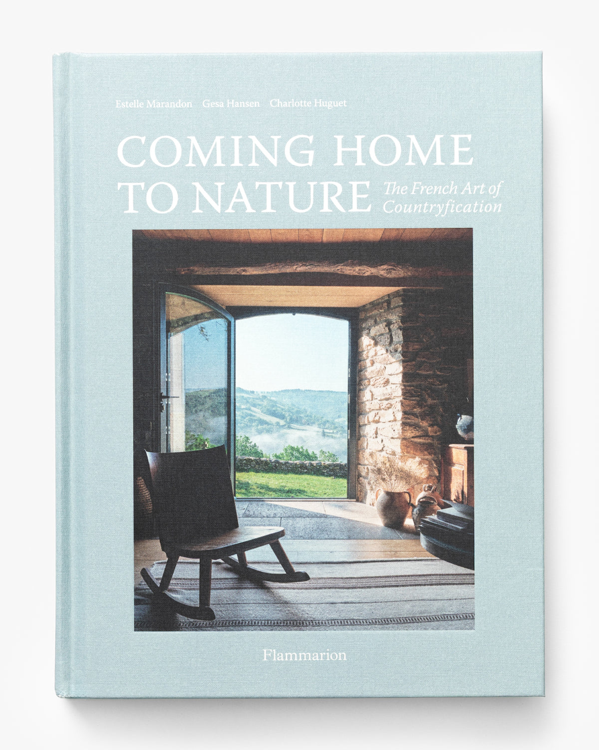 The cover of Coming Home to Nature: The French Art of Countryfication shows an open doorway to the French countryside, a chair, and a basket inside a rustic stone room—an ideal addition to any coffee table book collection, by McGee & Co.