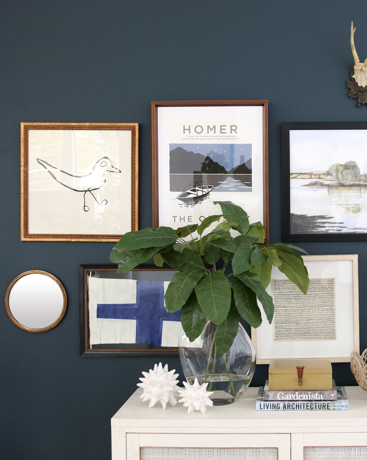 A sapphire blue wall displays framed art—one piece titled Repetition 7—above a white cabinet topped with a glass vase of green leaves, two spiky white sculptures, and stacked books.