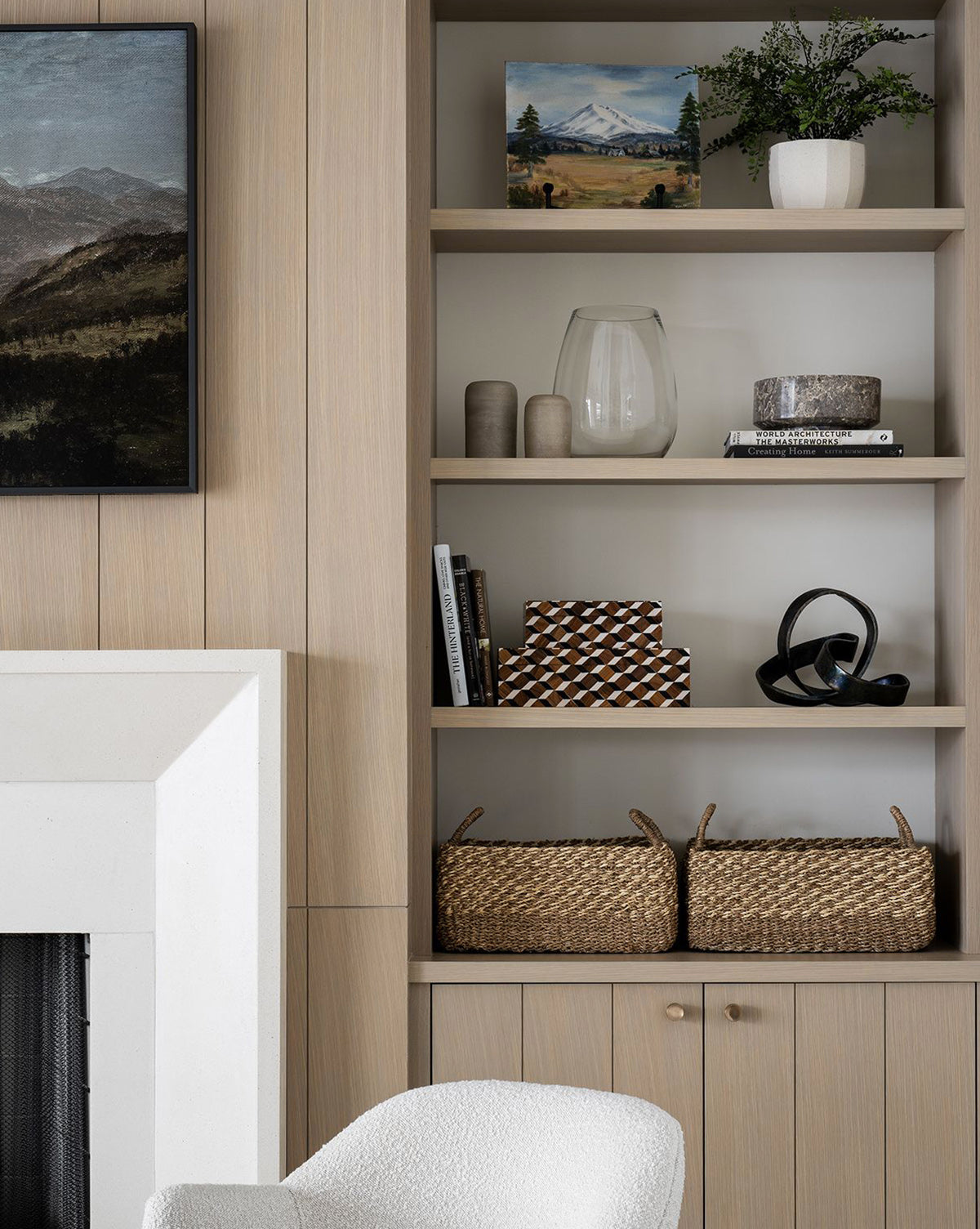 Built-in wooden shelves display decor, books, vases, a plant, framed art, and a Cordova Basket; a white fireplace and textured chair add to the cozy, modern living space.