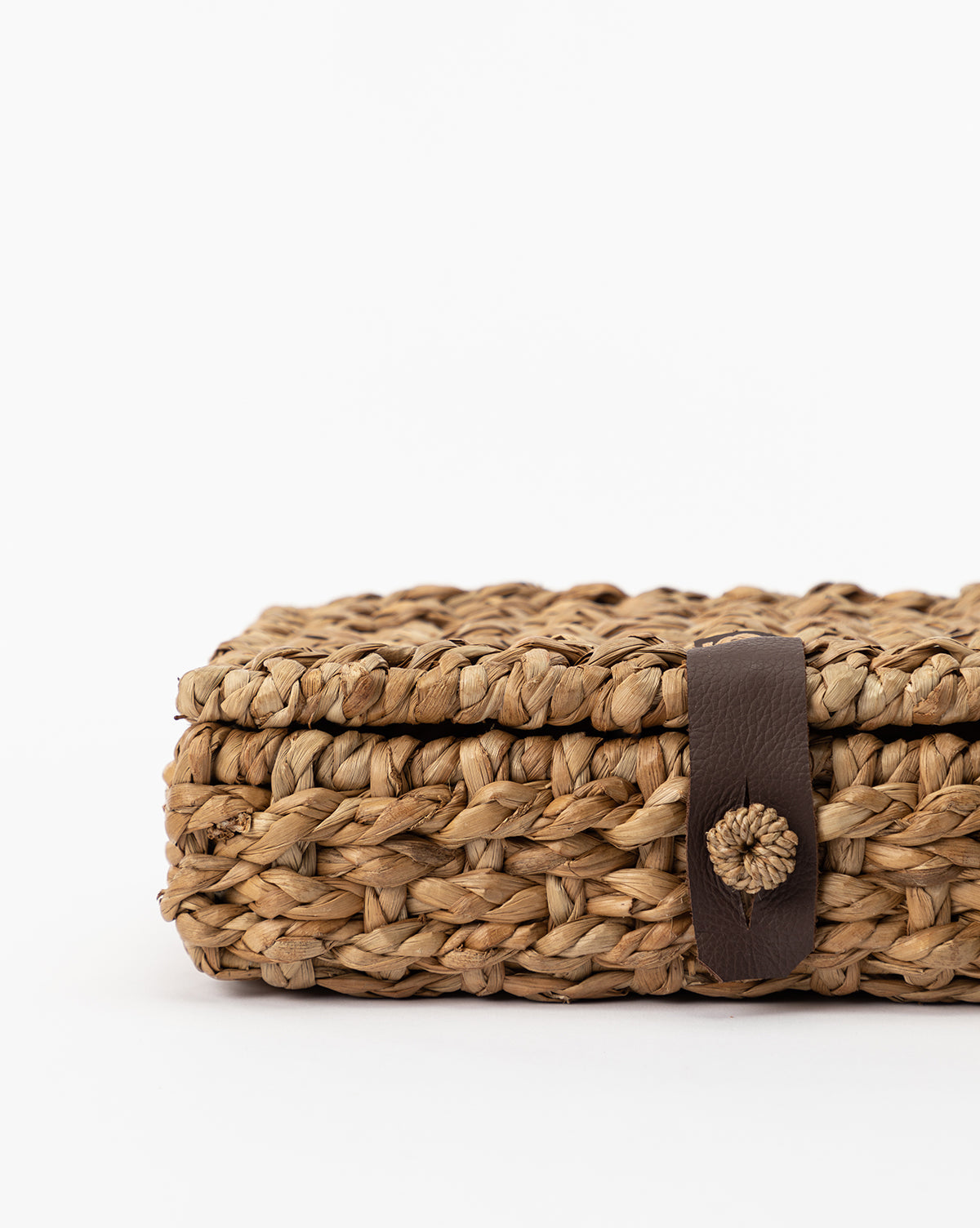 A close-up of the Corinne Woven Box—rectangular, crafted from natural seagrass fibers with a brown leather strap closure—displayed against a plain white background.