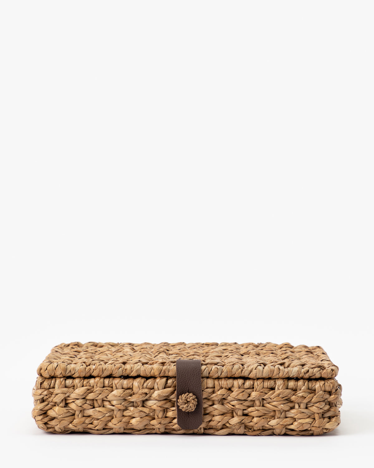 The Corinne Woven Box is a rectangular, natural seagrass storage piece with a lid, featuring a central brown strap and button closure. It’s photographed against a plain white background.