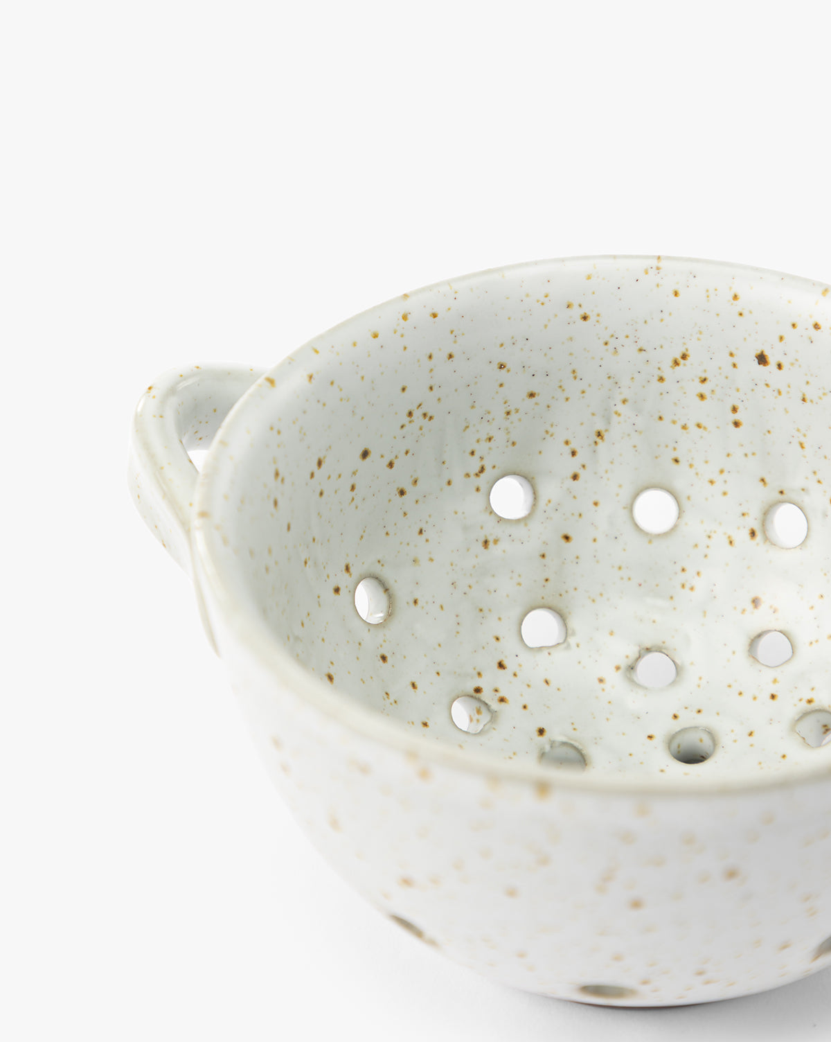 A close-up of the Cream Handled Berry Bowl, an artisanal stoneware piece with a small handle and multiple round holes in the bottom, set against a white background, by McGee & Co.