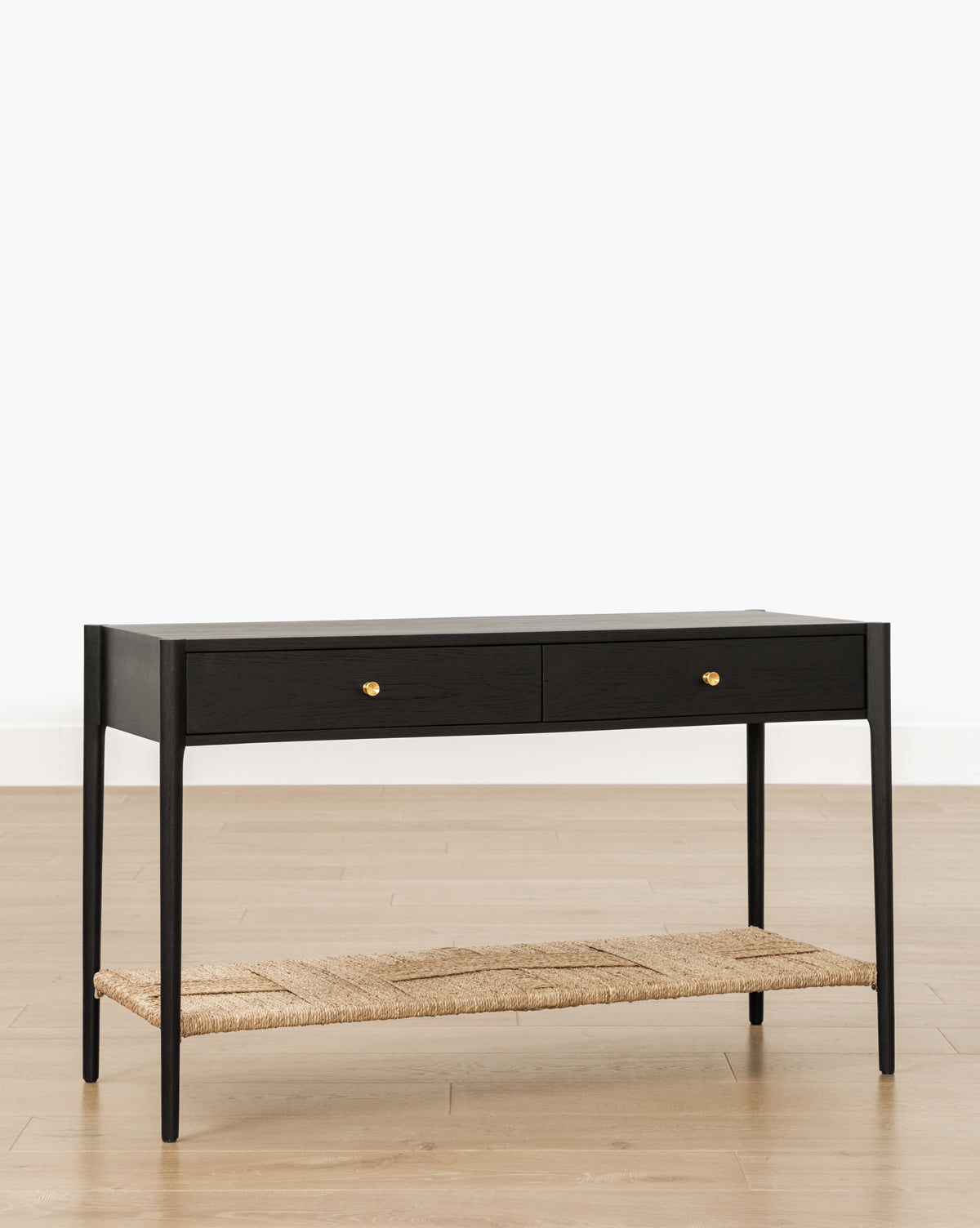 The Dana 2-Drawer Console, a modern black table with gold knobs and a woven lower shelf, is perfect for entryways, by McGee & Co. It stands on light wood flooring against a plain white background.