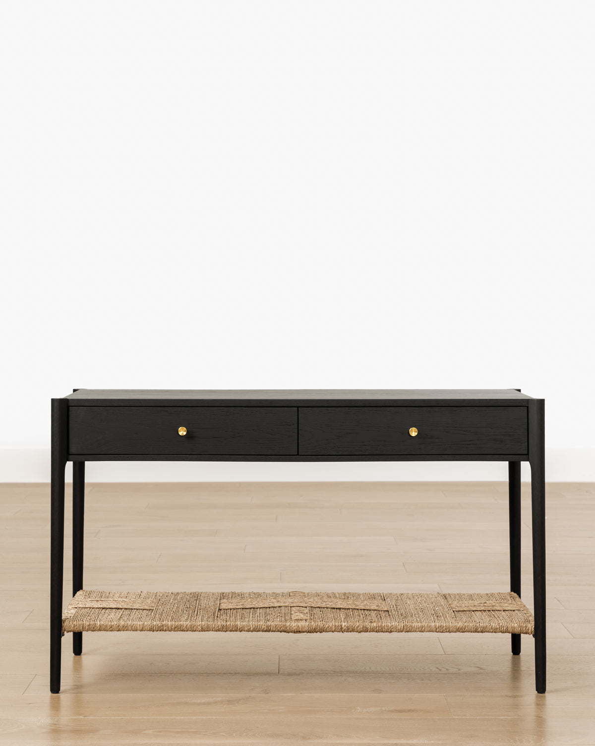 The Dana 2-Drawer Console features a modern black oak finish, gold knobs, and a woven rattan lower shelf. Standing on slender legs, its ideal for entryway decor against a white background on a light wood floor, by McGee & Co.