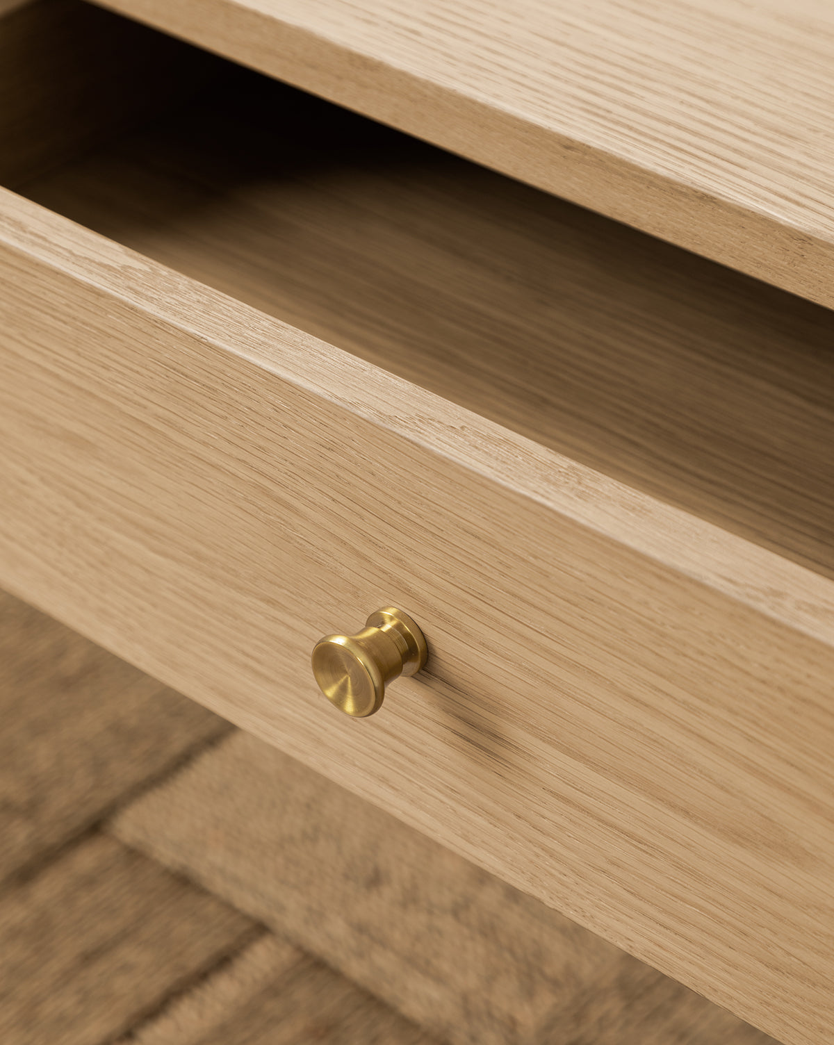 Close-up of a light wood drawer with a round brass knob on the Dana 2-Drawer Console, partially open to show the empty interior. This oak console table is ideal for entryway decor and sits on a textured surface by McGee & Co.