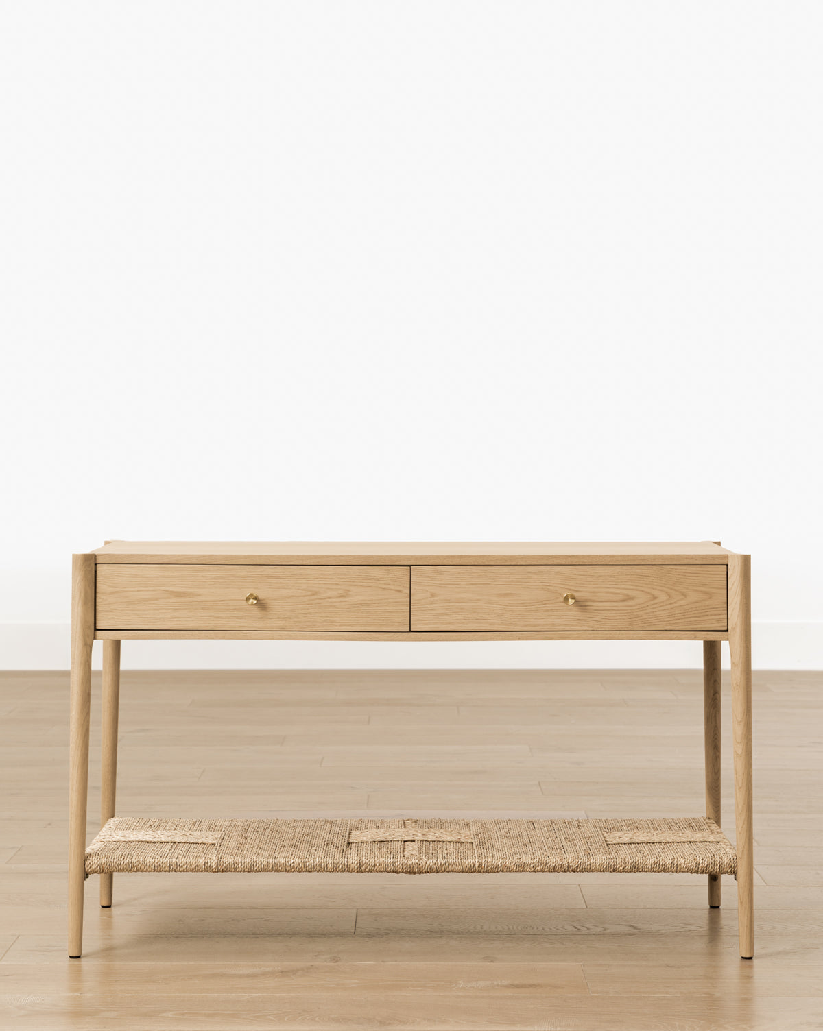 The Dana 2-Drawer Console features brass knobs and a woven lower shelf, ideal as an entryway accent, by McGee & Co. It stands on a light wood floor with a simple white background, offering both style and storage.