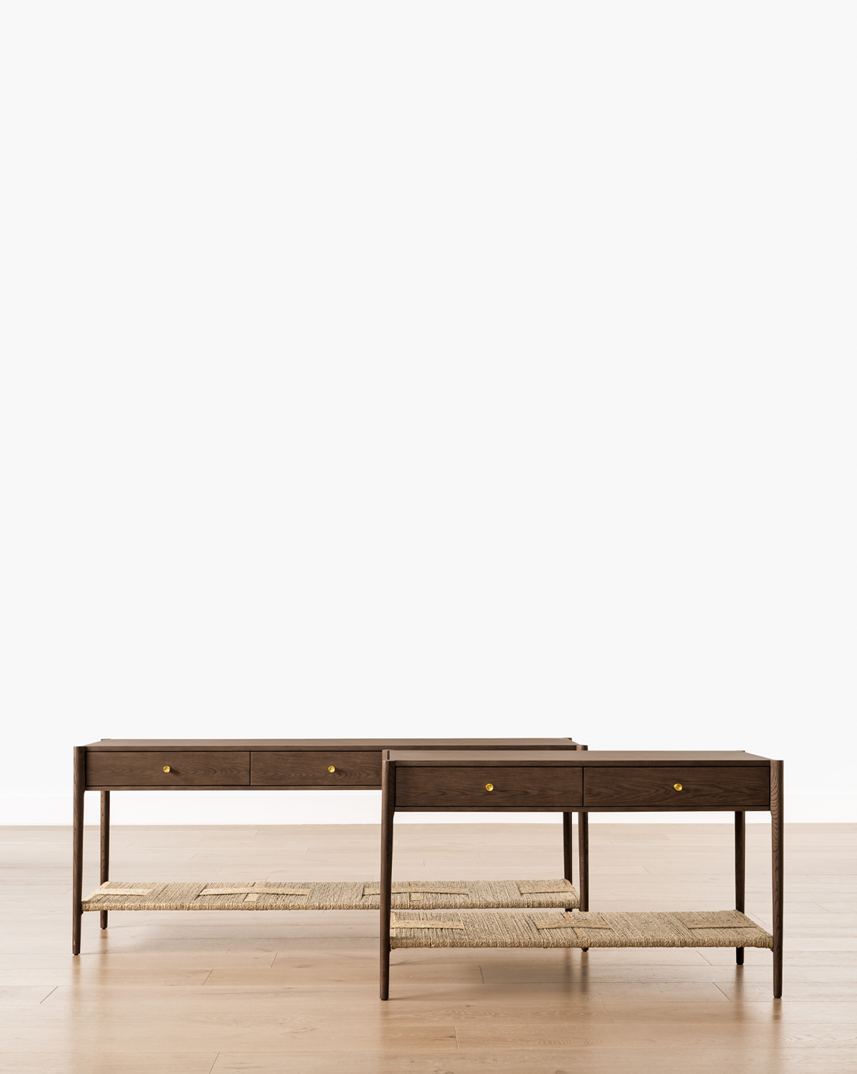 Two Dana Walnut Oak Console tables with woven shelves, small drawers, and brass hardware are placed side by side on a light wood floor against a plain white background, by McGee & Co.