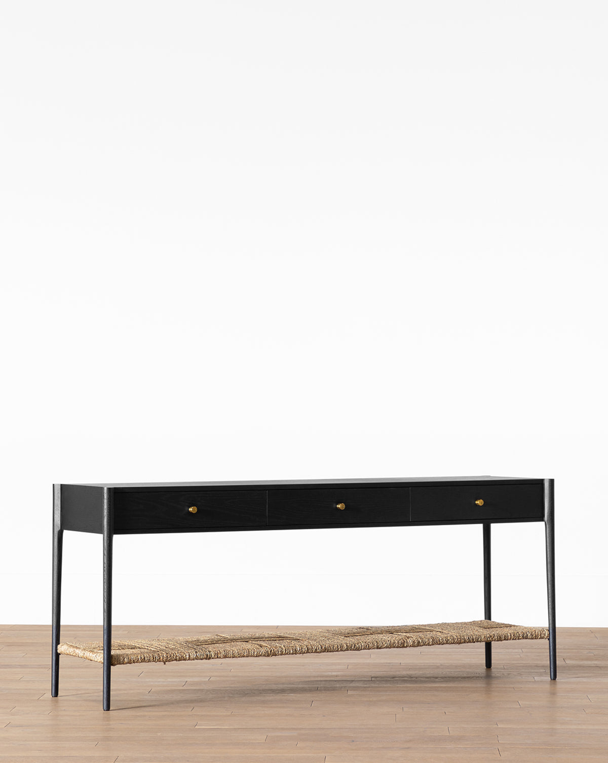 The Dana Console, a modern black entryway table with three small drawers, brass hardware, and a woven rattan lower shelf, stands on a wooden floor against a plain white background by McGee & Co.