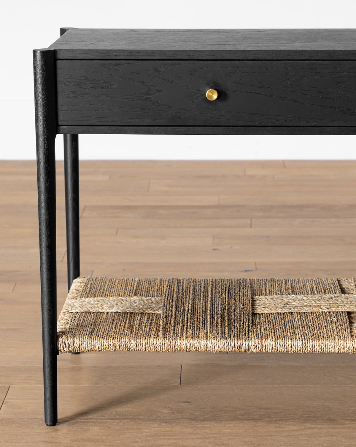 The Dana Console is a black oak table with a single brass-handled drawer and a woven natural fiber shelf, ideal for adding stylish entryway decor to a light wood floor by McGee & Co.