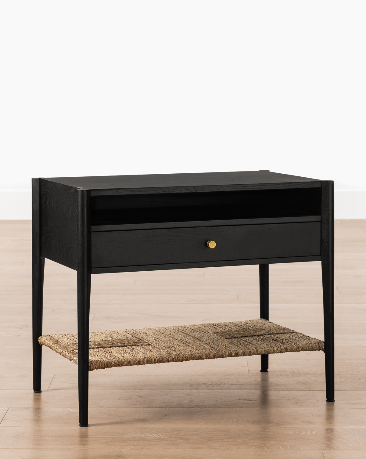 The Dana Nightstand features a sleek black finish, slim legs, a central drawer with gold knob, an open shelf above, and a woven seagrass shelf at the bottom, all resting on a light wood floor, by McGee & Co.