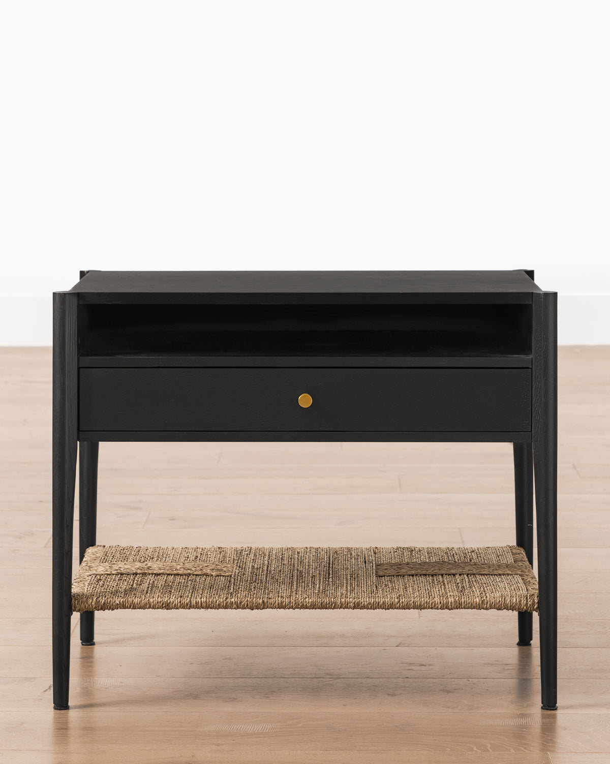 The Dana Nightstand features a modern black finish, single drawer with brass knob, open shelf above, and woven seagrass shelf below, shown on a light wood floor against a plain white background by McGee & Co.
