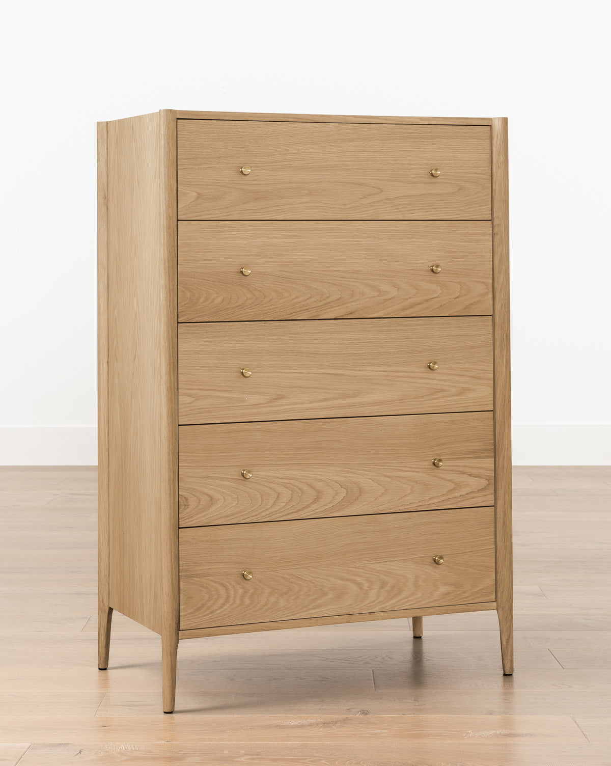 The Dana Tallboy Dresser, crafted from light white oak, has five drawers with round brass pulls and stands on four tapered legs. Its shown against a white wall and wooden floor by McGee & Co.