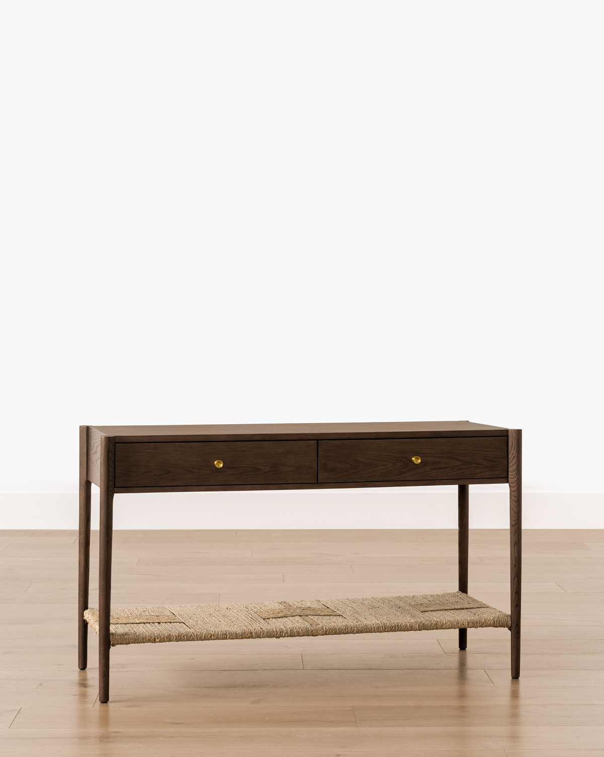 The Dana 2-Drawer Console, with brass knobs and a woven lower shelf in dark oak, brings timeless charm to your entryway when placed against a white wall and light wood floor, by McGee & Co.