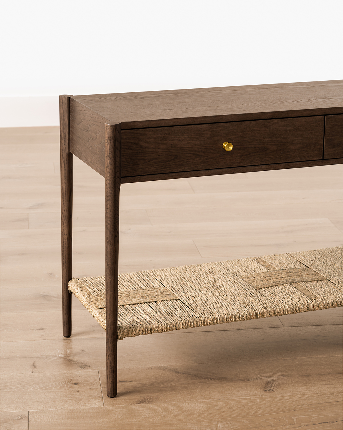 The Dana 2-Drawer Console features slim legs, brass knobs, and a woven rattan shelf to add charm to your entryway decor, set on a light wood floor against a white wall, by McGee & Co.